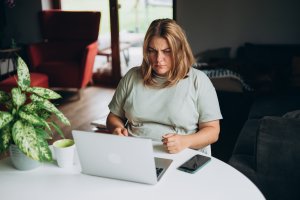 A woman with blonde hair sits at a table, looking concerned at her laptop screen as she reviews a lead. A smartphone, a cup, and a potted plant are on the table in a cozy, sunlit room.
