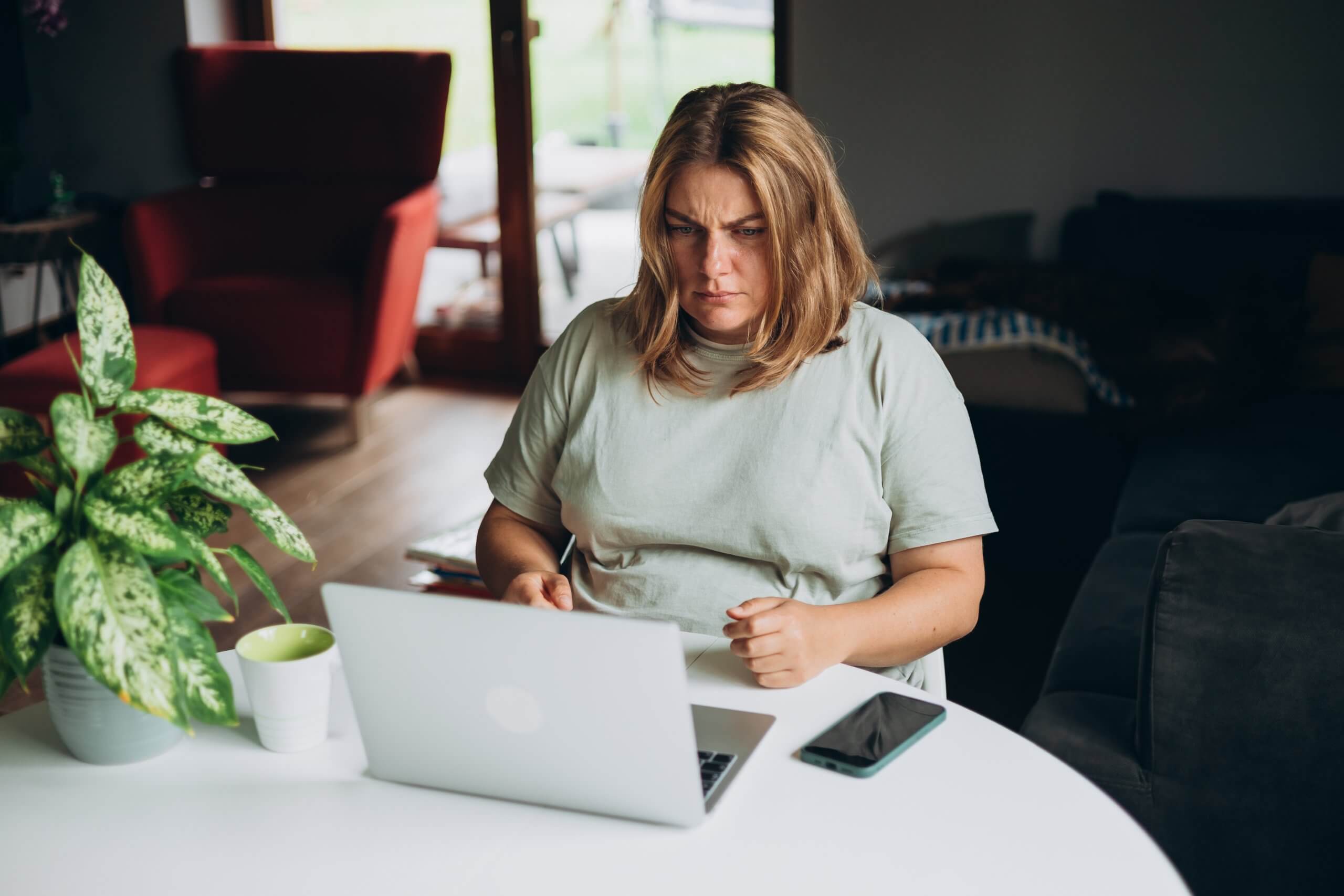 A woman with blonde hair sits at a table, looking concerned at her laptop screen as she reviews a lead. A smartphone, a cup, and a potted plant are on the table in a cozy, sunlit room. A woman with blonde hair sits at a table, looking concerned at her laptop screen as she reviews a lead. A smartphone, a cup, and a potted plant are on the table in a cozy, sunlit room.