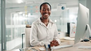 A woman in a white blouse sits at a desk in a modern office, smiling confidently at the camera. Surrounded by office supplies and technology, she embodies the professionalism and poise of today's leaders. Glass walls and shelves line the background.