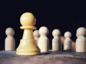 A yellow chess pawn stands prominently in the foreground on a wooden surface, symbolizing leadership amid several out-of-focus, beige wooden game pieces in the background against a dark backdrop.