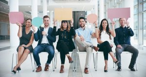 Six smiling people sit in a row on chairs in a bright, modern office, each holding up a colorful blank speech bubble over their heads, suggesting communication or sharing ideas—perfect for illustrating government discussions.