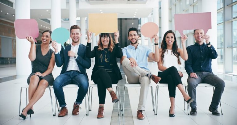 Six smiling people sit in a row on chairs in a bright, modern office, each holding up a colorful blank speech bubble over their heads, suggesting communication or sharing ideas—perfect for illustrating government discussions.