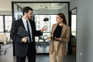 A man in a suit holding a folder and a woman in a tan suit holding a coffee cup are walking and having a conversation, discussing ways to lead their team in a modern office hallway.