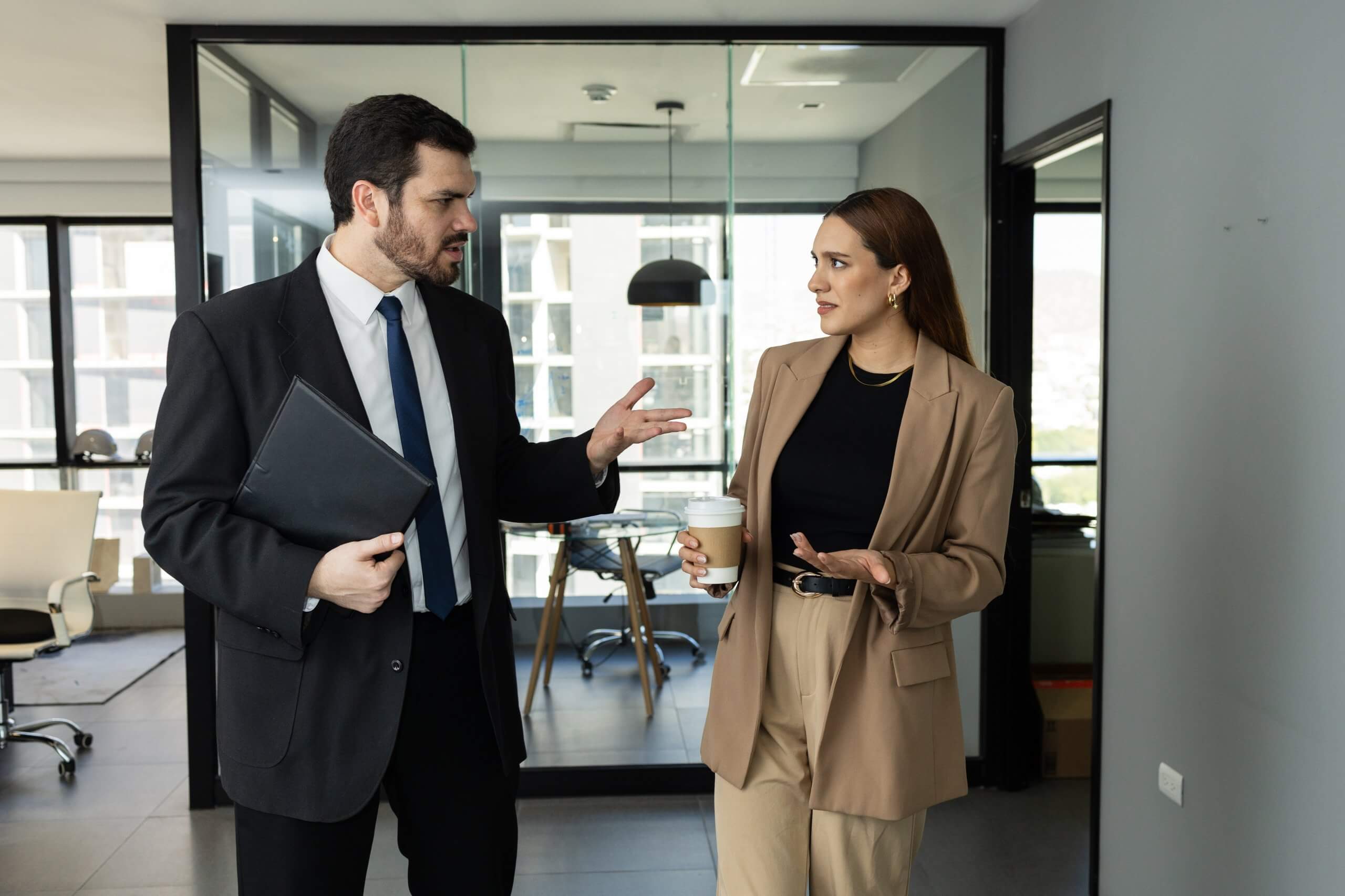 A man in a suit holding a folder and a woman in a tan suit holding a coffee cup are walking and having a conversation, discussing ways to lead their team in a modern office hallway. A man in a suit holding a folder and a woman in a tan suit holding a coffee cup are walking and having a conversation, discussing ways to lead their team in a modern office hallway.