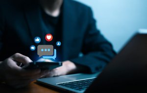 A person in a dark jacket uses a smartphone near an open laptop, with social media icons like thumbs up, heart, and speech bubble digitally floating above the phone—reflecting modern communication in the Public Sector.