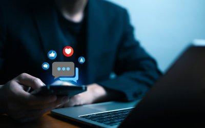 A person in a dark jacket uses a smartphone near an open laptop, with social media icons like thumbs up, heart, and speech bubble digitally floating above the phone—reflecting modern communication in the Public Sector.