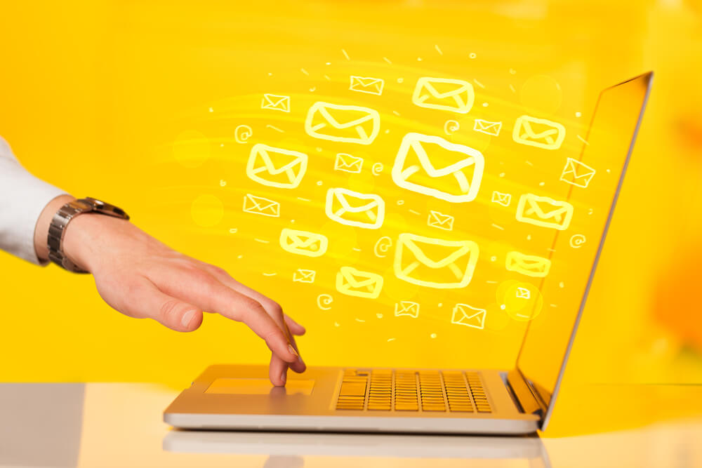 A hand reaching for a laptop keyboard, with illustrated yellow email icons—symbolizing automation—appearing to come out of the screen on a bright yellow background.