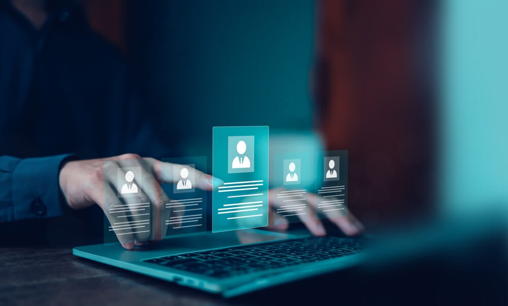 A person uses a laptop, with digital profile icons and information cards appearing above the keyboard, suggesting a virtual recruitment process. A person uses a laptop, with digital profile icons and information cards appearing above the keyboard, suggesting a virtual recruitment process.