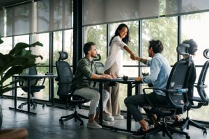 Three people in a modern office, two sitting at desks and one standing, smiling and shaking hands across the table. Large windows and greenery are visible in the background, reflecting the collaborative spirit often found in public agencies.
