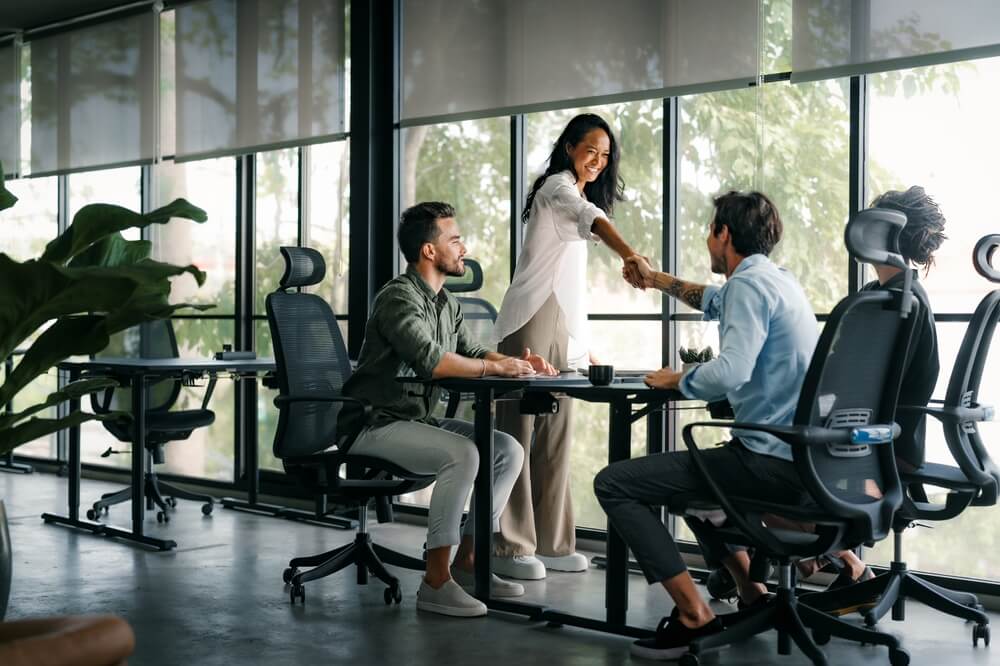 Three people in a modern office, two sitting at desks and one standing, smiling and shaking hands across the table. Large windows and greenery are visible in the background, reflecting the collaborative spirit often found in public agencies.