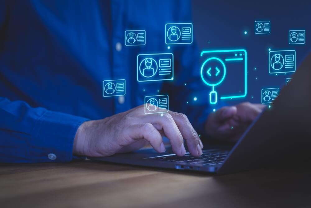 A person types on a laptop with digital icons of user profiles, a magnifying glass, and code brackets hovering above the keyboard, representing concepts of recruitment, user search, coding, and data management. A person types on a laptop with digital icons of user profiles, a magnifying glass, and code brackets hovering above the keyboard, representing concepts of recruitment, user search, coding, and data management.