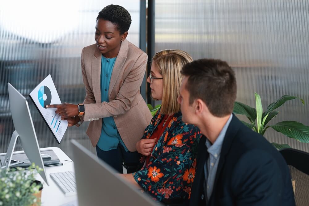 Three colleagues in an office sit at computers while a woman stands, pointing to a pie chart on paper, explaining workforce data to the others. Large windows and plants are in the background. Three colleagues in an office sit at computers while a woman stands, pointing to a pie chart on paper, explaining workforce data to the others. Large windows and plants are in the background.
