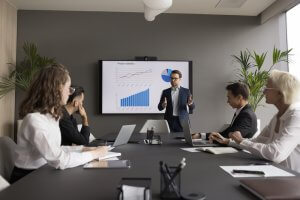 A man in a suit presents project statistics on a screen to four workers seated at a conference table with laptops, notebooks, and papers in a modern office meeting room with plants.