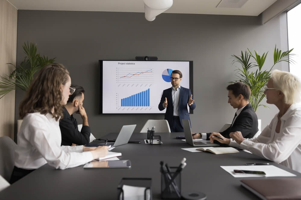 A man in a suit presents project statistics on a screen to four workers seated at a conference table with laptops, notebooks, and papers in a modern office meeting room with plants. A man in a suit presents project statistics on a screen to four workers seated at a conference table with laptops, notebooks, and papers in a modern office meeting room with plants.