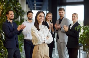 A confident woman stands smiling with arms crossed in a modern office, while five coworkers from public agencies clap and smile behind her, surrounded by green plants and stylish decor.
