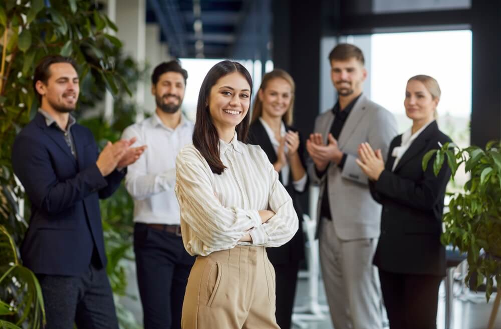 A confident woman stands smiling with arms crossed in a modern office, while five coworkers from public agencies clap and smile behind her, surrounded by green plants and stylish decor.