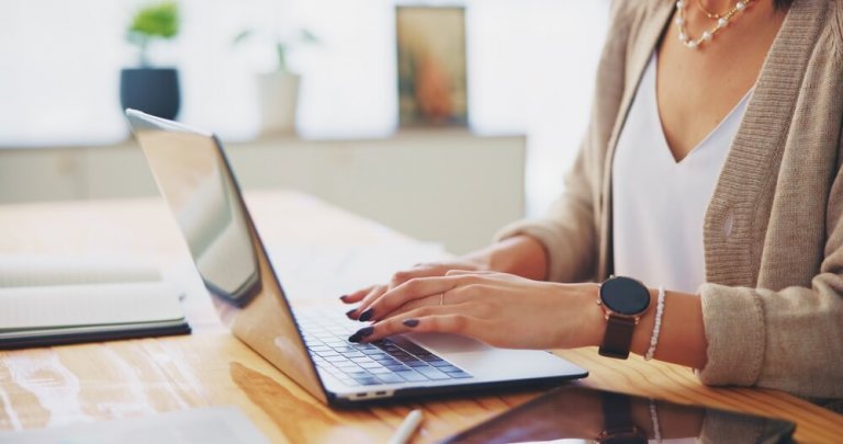 A woman wearing a blazer and jewelry types on a laptop at a wooden desk, exploring automation tools, with a notebook, tablet, and blurred office background visible.