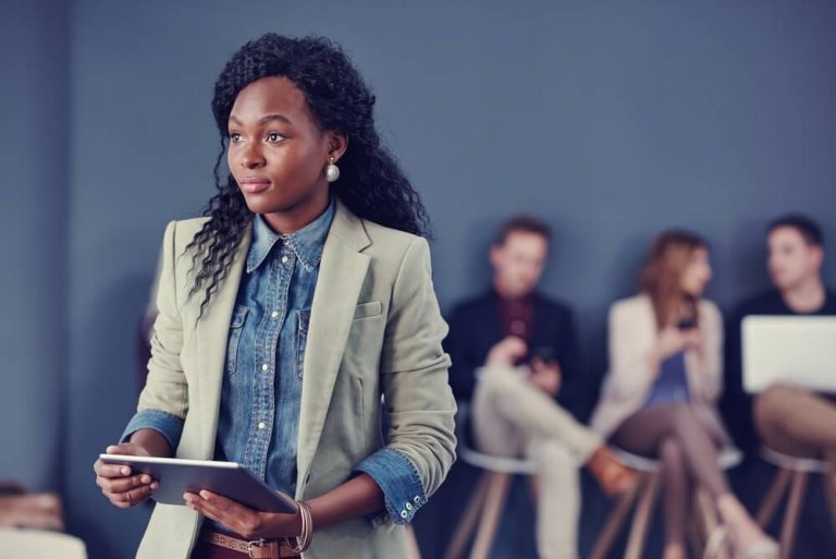 A woman in a blazer and denim shirt holds a tablet and looks to the side, standing in front of a blurred group involved in a recruitment discussion.