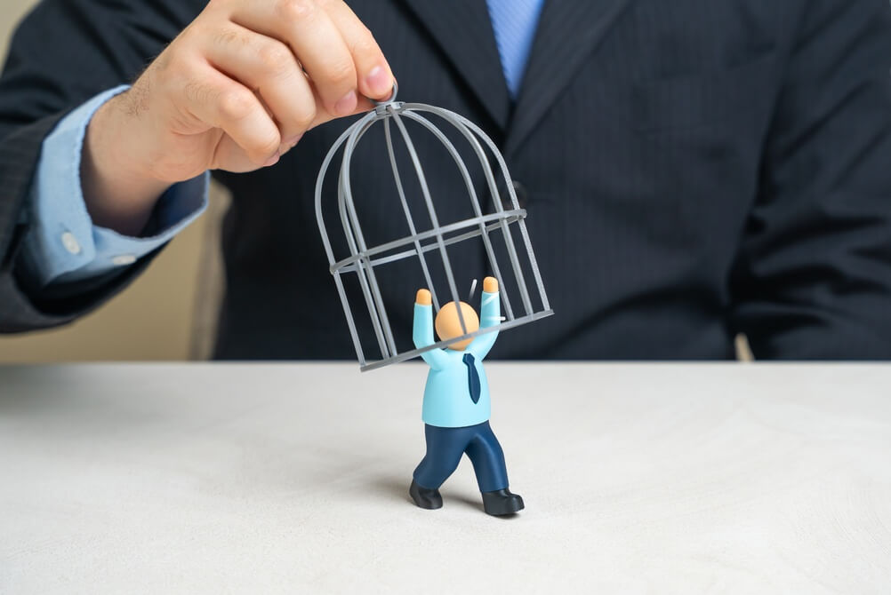 A person in a suit lifts a wire cage off a small figurine dressed in business attire, symbolizing freedom or release from confinement in the context of recruitment, on a white surface. A person in a suit lifts a wire cage off a small figurine dressed in business attire, symbolizing freedom or release from confinement in the context of recruitment, on a white surface.