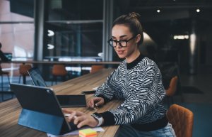A woman wearing glasses and a zebra-print sweater works on a tablet at a long wooden table in a modern, brightly lit office space that reflects a vibrant and collaborative culture. Colorful sticky notes are on the table near her hand.