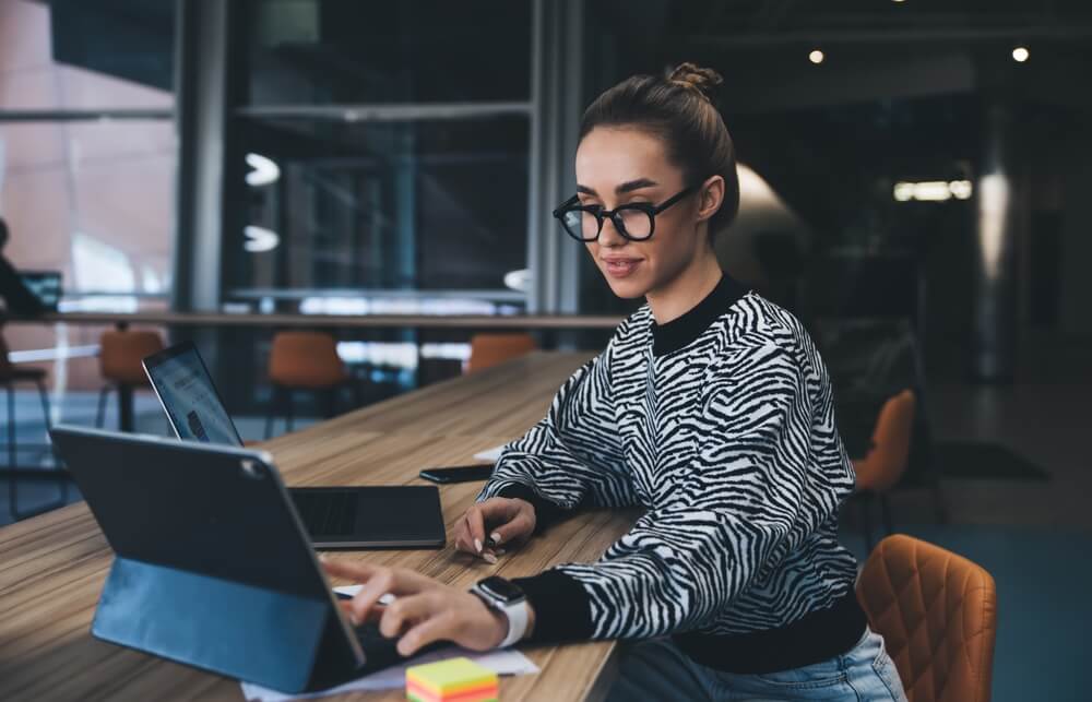 A woman wearing glasses and a zebra-print sweater works on a tablet at a long wooden table in a modern, brightly lit office space that reflects a vibrant and collaborative culture. Colorful sticky notes are on the table near her hand.