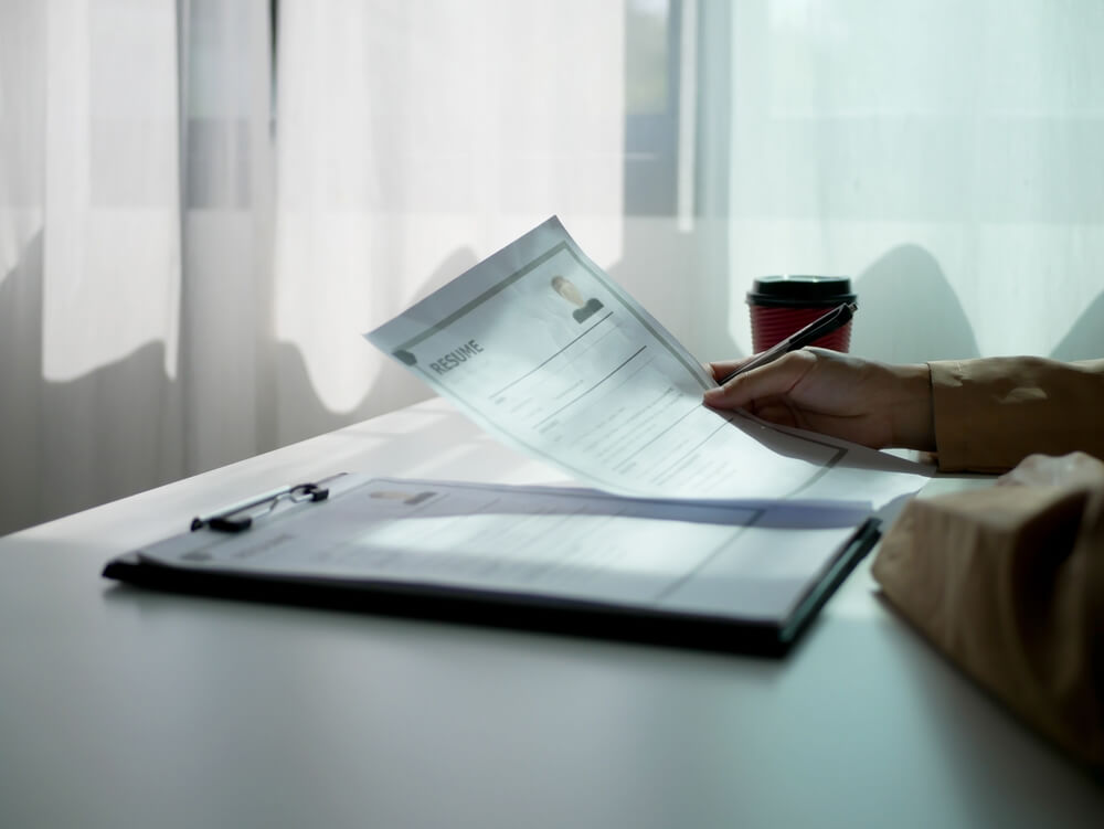 A person reviews a printed resume at a desk, with sunlight streaming through sheer curtains in the background and a red coffee cup nearby, thoughtfully considering qualifications for a government position. A person reviews a printed resume at a desk, with sunlight streaming through sheer curtains in the background and a red coffee cup nearby, thoughtfully considering qualifications for a government position.