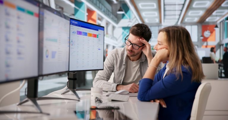 Two people sit at a modern office desk, looking intently at computer monitors displaying colorful data charts and graphs in an open, bright workspace, analyzing key recruitment metrics to drive hiring strategies.
