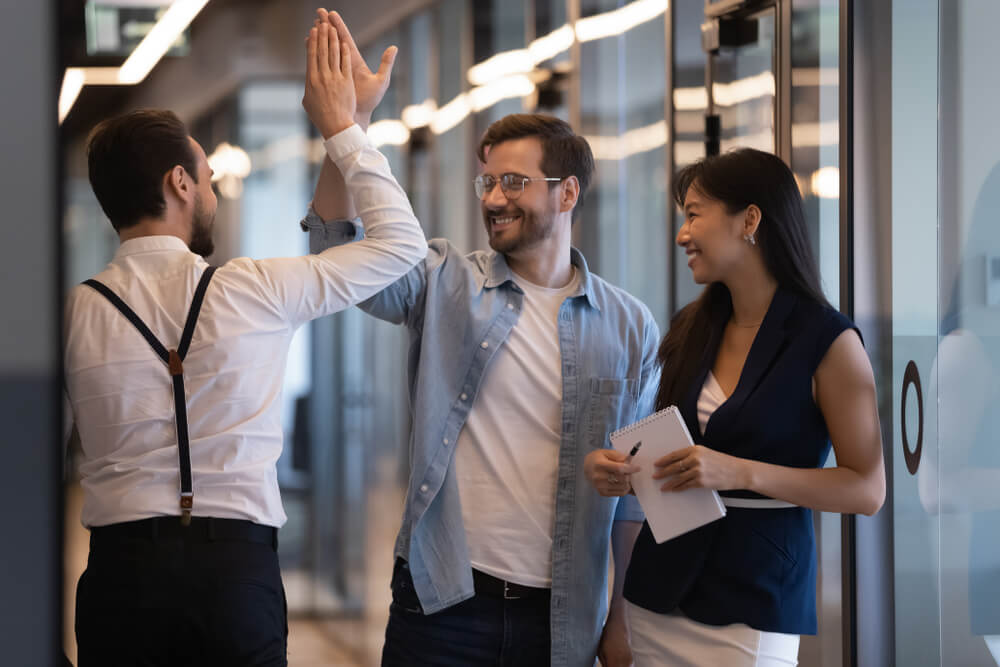 Three coworkers stand in an office hallway, embodying a positive workplace culture; two men are giving each other a high-five while a woman next to them smiles and holds a notepad.
