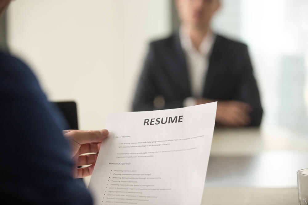 A person holds a resume during a government job interview, with another person in a suit sitting across the desk, slightly out of focus. A person holds a resume during a government job interview, with another person in a suit sitting across the desk, slightly out of focus.