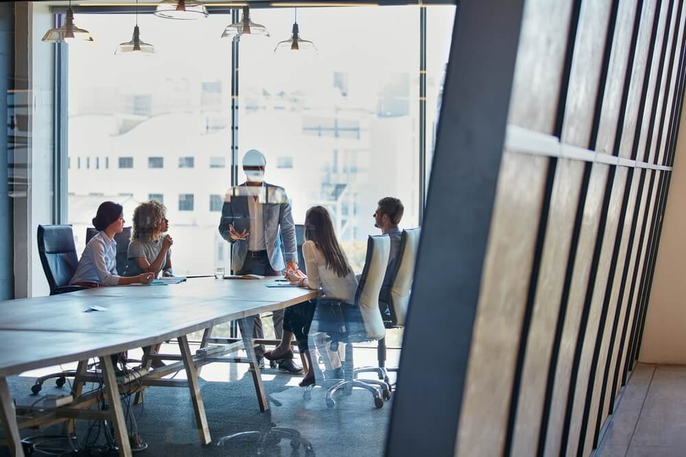 Five team members in business attire sit around a conference table in a modern office, with one person standing and presenting near large windows letting in natural light.
