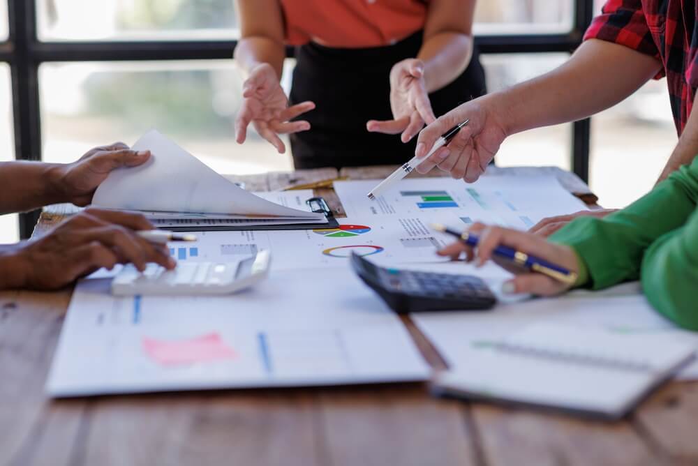 Four people discuss documents and charts on a wooden table, using calculators, pens, and papers with graphs and data. Their collaborative communication is evident as only their hands and arms are visible while they work together in a bright office room. Four people discuss documents and charts on a wooden table, using calculators, pens, and papers with graphs and data. Their collaborative communication is evident as only their hands and arms are visible while they work together in a bright office room.