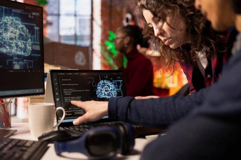 Two government workers collaborate on a laptop displaying code, with a digital brain graphic on a monitor nearby. A coffee mug and headphones sit on the desk; another person works in the background.