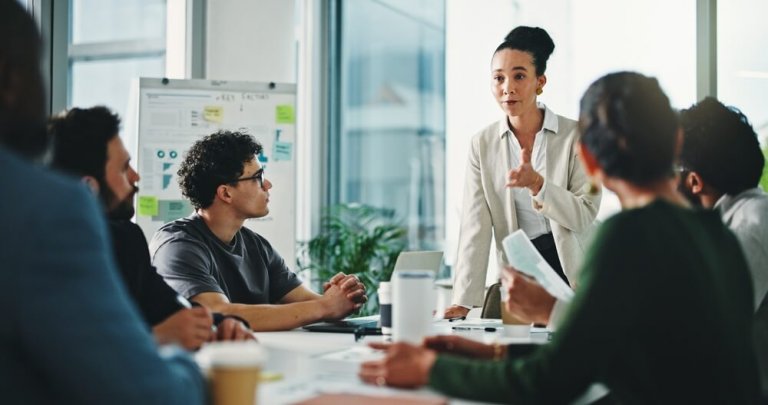 A woman stands and speaks to her team of colleagues seated around a conference table during a meeting, with charts and papers visible in the background.