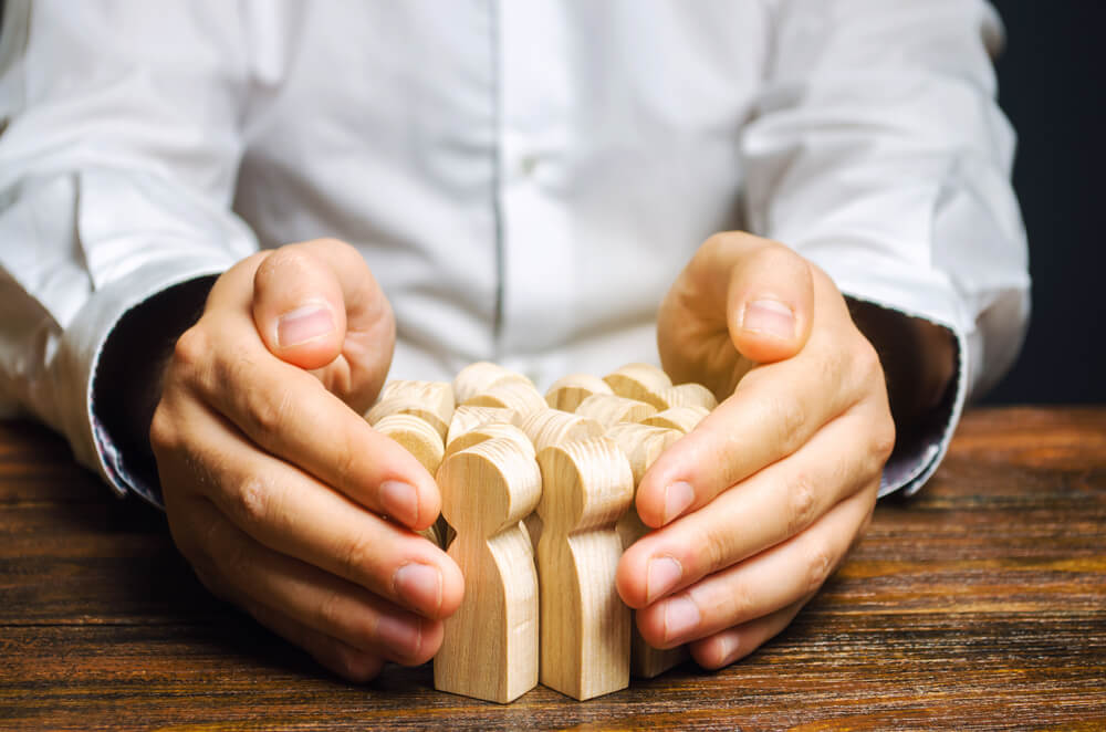 A person in a white shirt cups their hands protectively around a group of small wooden human figures on a wooden table, symbolizing the care and responsibility involved in recruitment. A person in a white shirt cups their hands protectively around a group of small wooden human figures on a wooden table, symbolizing the care and responsibility involved in recruitment.