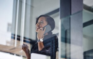 A woman smiles while talking on her smartphone, standing indoors near large glass windows. Engaged in cheerful communication, she is wearing a black top and a wristwatch, and appears happy, possibly celebrating good news.