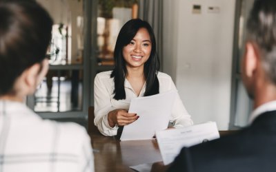A woman in a white blouse smiles while holding papers during a government meeting or interview with two people, seen from behind, across a table in an office setting.