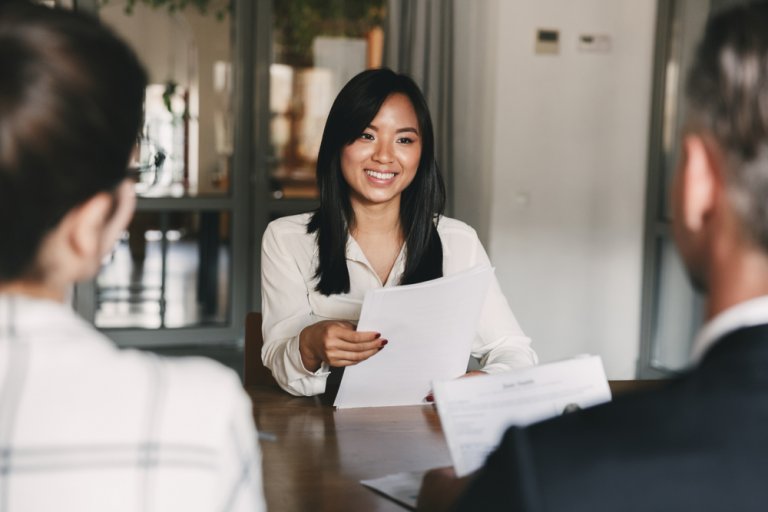 A woman in a white blouse smiles while holding papers during a government meeting or interview with two people, seen from behind, across a table in an office setting.