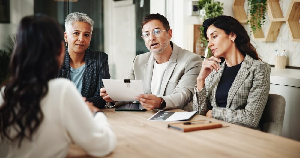 Interview,,Cv,And,Business,People,In,Office,For,Recruitment,Process Five team members in business attire sit around a conference table in a modern office, with one person standing and presenting near large windows letting in natural light.