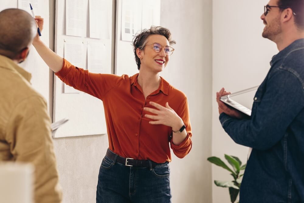 Two men in casual clothes smile and look at a tablet together in a modern office space, highlighting collaboration, while a woman in the background stands near desks and computers.