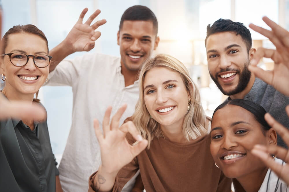 Selfie,,Smile,And,Diversity,At,Office,With,Ok,Hand,Sign, A woman wearing glasses and a zebra-print sweater works on a tablet at a long wooden table in a modern, brightly lit office space that reflects a vibrant and collaborative culture. Colorful sticky notes are on the table near her hand.