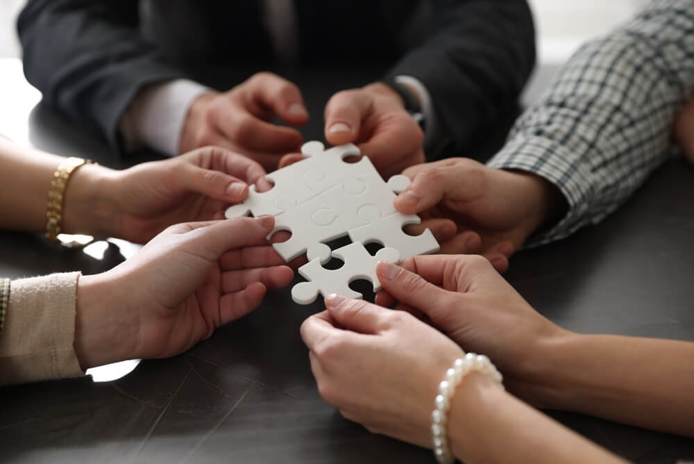 Team,Building.,Group,Of,Businesspeople,Putting,Puzzle,Pieces,Together,At A man in a red shirt sits at a desk with a laptop, demonstrating excellent communication skills as he talks on the phone and holds a clipboard, smiling.