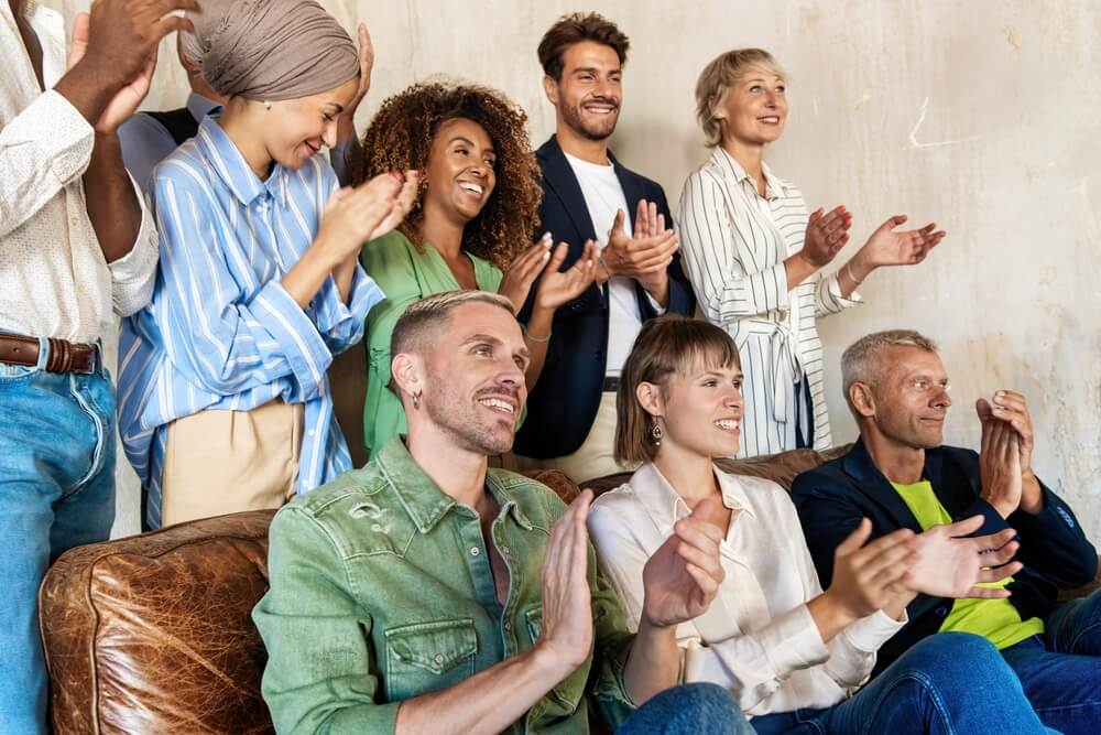 Enthusiastic,Diverse,Team,Clapping,And,Cheering.,Multicultural,,Multi-generational,Group,Showing Two men in casual clothes smile and look at a tablet together in a modern office space, highlighting collaboration, while a woman in the background stands near desks and computers.