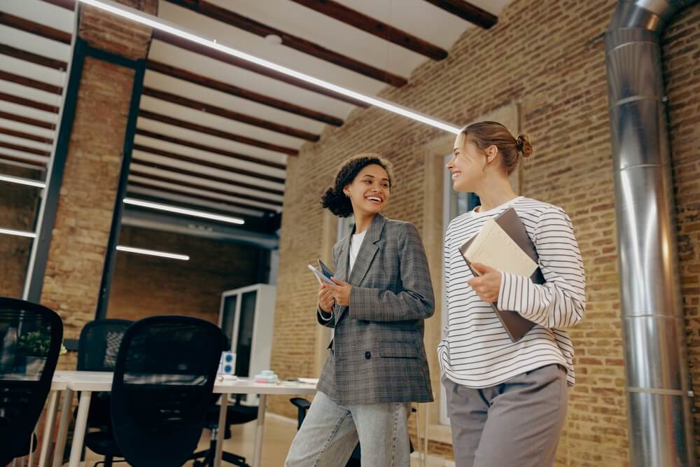 Smiling,Women,Colleagues,Speak,Laughing,Enjoy,Leisure,Time,At,Workplace. Three coworkers stand in an office hallway, embodying a positive workplace culture; two men are giving each other a high-five while a woman next to them smiles and holds a notepad.