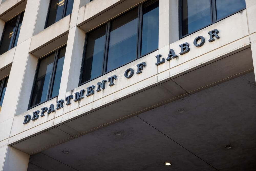 Signage,Outside,Of,The,United,States,Department,Of,Labor,Headquarters The United States flag waves in front of a large federal government building with tall white columns and a blue sky in the background.