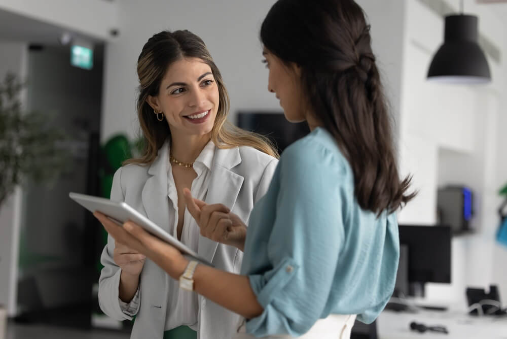 Young,Businesswoman,Listening,To,Experienced,Coworker,With,Digital,Tablet,Receiving A person in a white shirt cups their hands protectively around a group of small wooden human figures on a wooden table, symbolizing the care and responsibility involved in recruitment.
