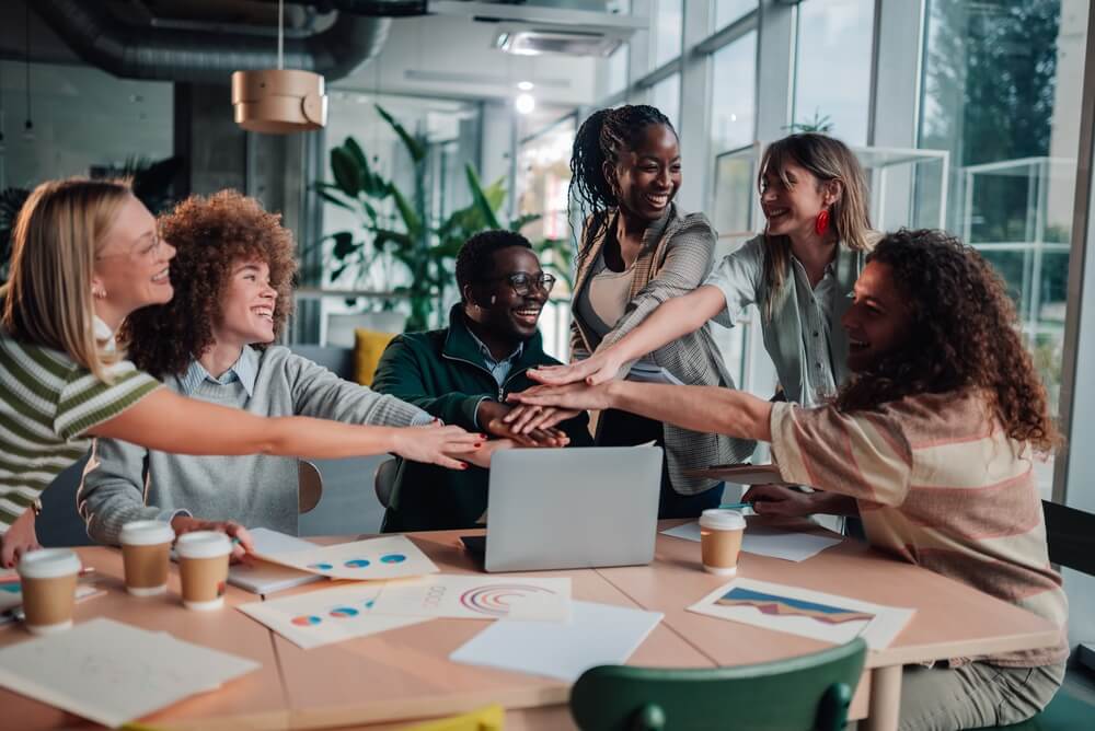 Happy,Business,Team,Members,Stacking,Hands,Together,In,A,Modern Four people discuss documents and charts on a wooden table, using calculators, pens, and papers with graphs and data. Their collaborative communication is evident as only their hands and arms are visible while they work together in a bright office room.