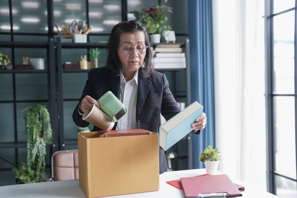 Quit,Job,Senior,Asian,Business,Woman,Sending,Resignation,Letter,And Five team members in business attire sit around a conference table in a modern office, with one person standing and presenting near large windows letting in natural light.