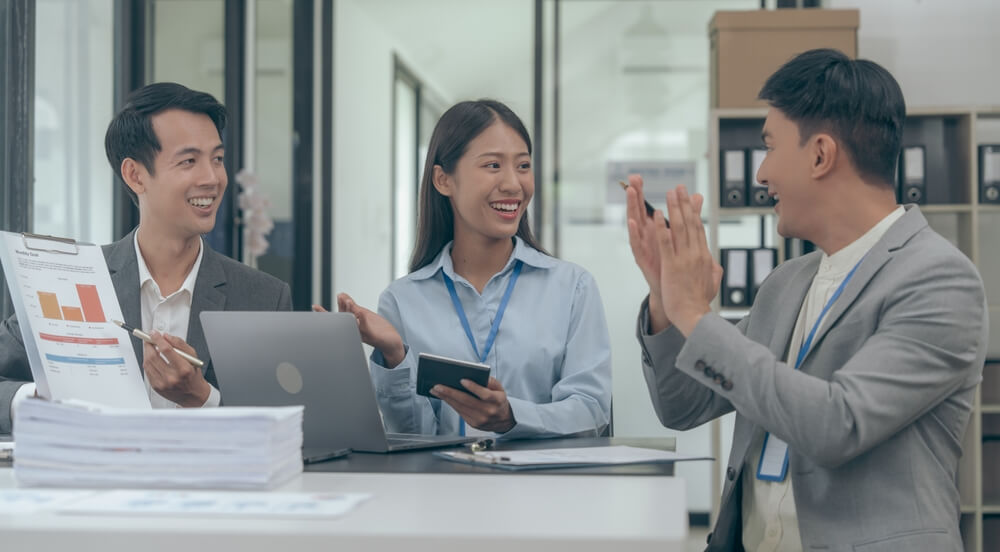 Three coworkers stand in an office hallway, embodying a positive workplace culture; two men are giving each other a high-five while a woman next to them smiles and holds a notepad.