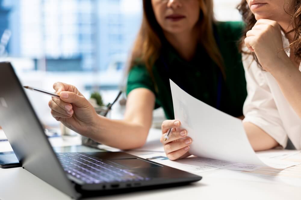 Hr,Coworkers,Using,Laptop,To,Do,Preemployment,Assessments,And,Background A person in a white shirt cups their hands protectively around a group of small wooden human figures on a wooden table, symbolizing the care and responsibility involved in recruitment.